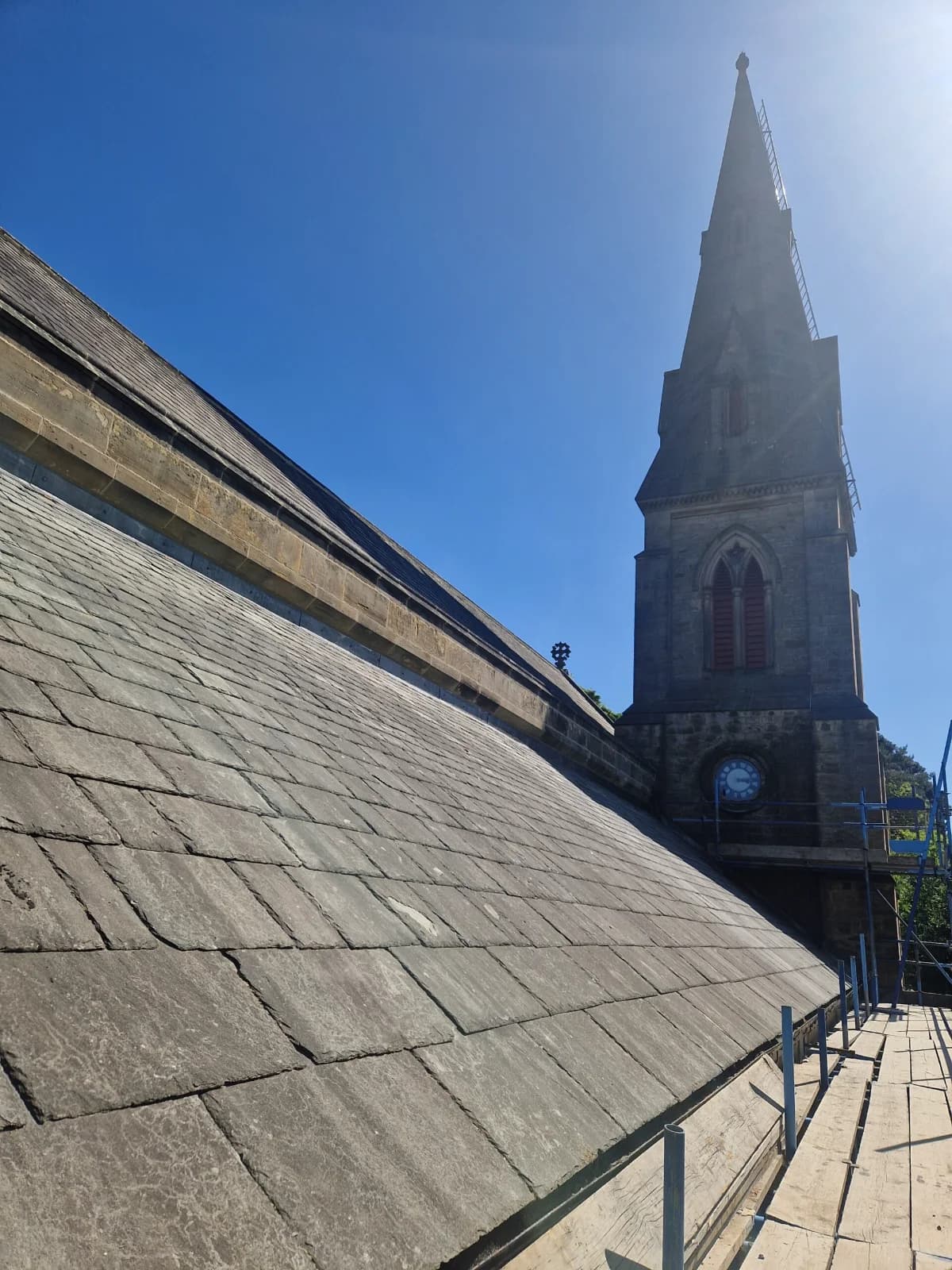 Stone gothic church mid-slate-roof works — spire and main roof in sunlight, scaffold visible — heritage roofing contractor County Durham