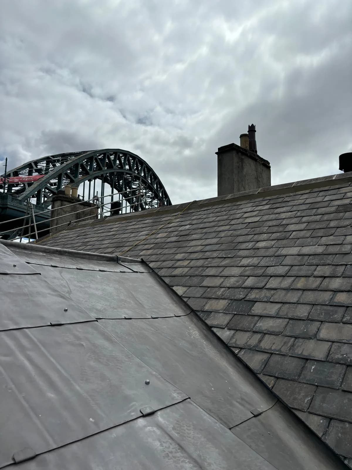 Completed natural slate roof with an adjacent lead-clad section and rebuilt brick chimney, the Tyne Bridge visible behind — Newcastle heritage roofing